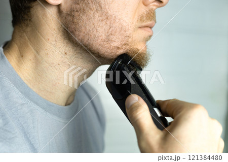 A man trims his beard with a trimmer. An electric razor trimming a beard. Beard care at home. A man trims his beard with a trimmer. An electric razor trimming a beard. Beard care at home. 121384480
