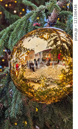 Shiny golden Christmas ornament reflecting festive market scene, surrounded by snow-covered evergreen branches and warm lights 121386436
