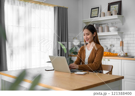 Beautiful young Asian woman working with laptop computer in home kitchen. Work at home Beautiful young Asian woman working with laptop computer in home kitchen. Work at home 121388400