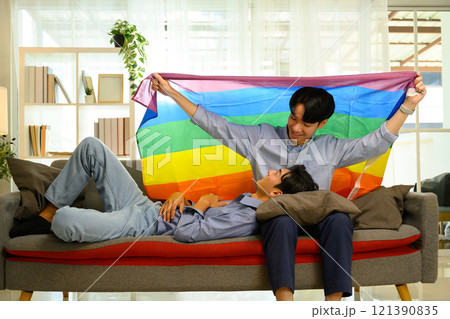 Cheerful gay couple holding a rainbow pride flag relaxing on sofa 121390835