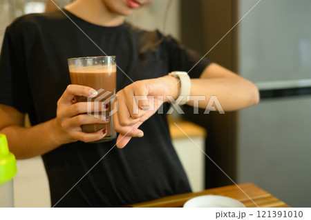 Young man holding a glass of chocolate protein shake and checking her smartwatch Young man holding a glass of chocolate protein shake and checking her smartwatch 121391010