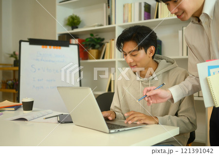 Asian male student in a hoodie working on a laptop, guided by a tutor at home 121391042