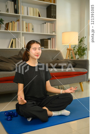 Young woman with eyes closed practicing meditation on a blue exercise mat in living room 121391085