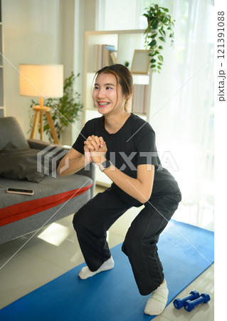 Beautiful young woman doing squat exercise on mat in her living room. Active and healthy lifestyle Beautiful young woman doing squat exercise on mat in her living room. Active and healthy lifestyle 121391088