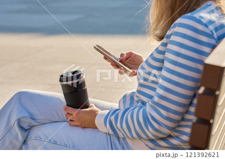 Woman using smartphone and holding reusable coffee cup while sitting on bench Woman using smartphone and holding reusable coffee cup while sitting on bench 121392621