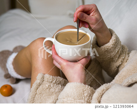 Woman in a bathrobe is sitting on a white bed, stirring a cup of coffee with a spoon. There are tangerines scattered on the bed around her 121393064