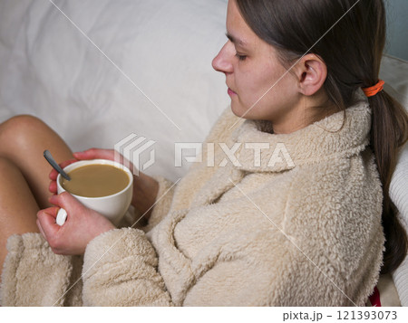 Relaxed young woman in comfortable bathrobe enjoying cozy moment with warm cup of coffee and on the sofa 121393073
