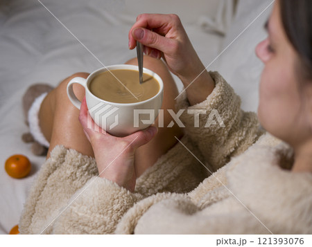 Young woman enjoys a cozy morning at home, wearing a beige bathrobe and stirring her coffee with a metal spoon, embracing relaxation during breakfast 121393076