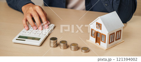 A person presses a calculator next to a house model and coin stacks, analyzing tax, property value, and mortgage payments. Highlights real estate profit, cash savings A person presses a calculator next to a house model and coin stacks, analyzing tax, property value, and mortgage payments. Highlights real estate profit, cash savings 121394002