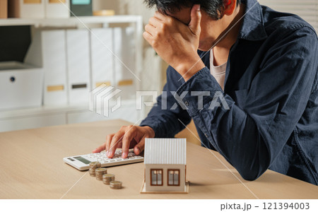 Stressed person holding a calculator, with stacks of coins and a house model nearby. Reflects financial planning challenges, tax concerns, and property calculations causing worry 121394003