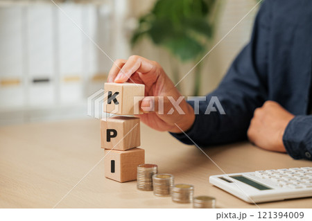 A close-up of a businessman hand holding wooden cube blocks spelling KPI near coins and a calculator. Represents KEY PERFORMANCE INDEX planning, employee performance tracking A close-up of a businessman hand holding wooden cube blocks spelling KPI near coins and a calculator. Represents KEY PERFORMANCE INDEX planning, employee performance tracking 121394009