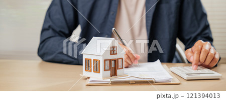 A man writes a signature on house insurance documents, using a calculator for tax and property details. Counting payments and rent by model house, he finalizes the price A man writes a signature on house insurance documents, using a calculator for tax and property details. Counting payments and rent by model house, he finalizes the price 121394013