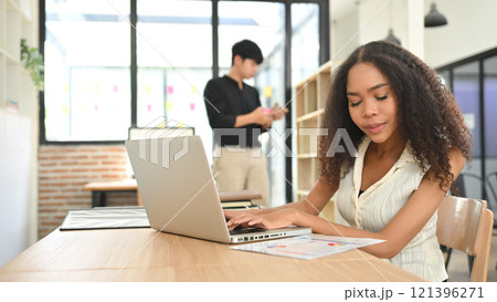 African American businesswoman reviewing charts and working on laptop at modern office desk African American businesswoman reviewing charts and working on laptop at modern office desk 121396271