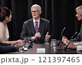 Senior politician in formal wear chairing committee answering questions during press conference while sitting at table with other administration officials 121397465