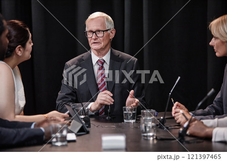 Senior politician in formal wear chairing committee answering questions during press conference while sitting at table with other administration officials Senior politician in formal wear chairing committee answering questions during press conference while sitting at table with other administration officials 121397465