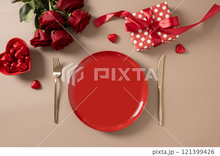 Valentine's day. Romantic dinner for date with red plate, chocolate sweets, romantic gift and red roses on beige background. View from above. Copy space. 121399426