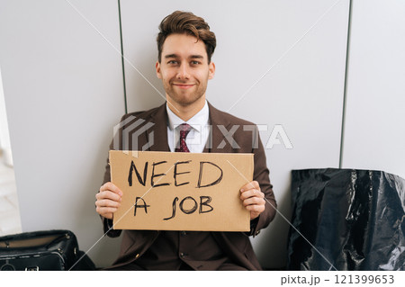 Portrait of cheerful businessman in suit holding cardboard sign with inscription need job, looking for work after being fired. Homeless young manager searching job sitting on urban street. Portrait of cheerful businessman in suit holding cardboard sign with inscription need job, looking for work after being fired. Homeless young manager searching job sitting on urban street. 121399653