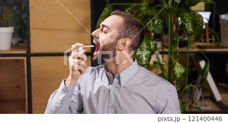 Young man feels with symptom of ill, sore throat ache and use spray to remove this pain sitting at desk at office. . 121400446
