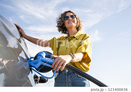 Woman with fueling hydrogen car. Refueling car with hydrogen fuel. Wind turbines in the background Woman with fueling hydrogen car. Refueling car with hydrogen fuel. Wind turbines in the background 121400636