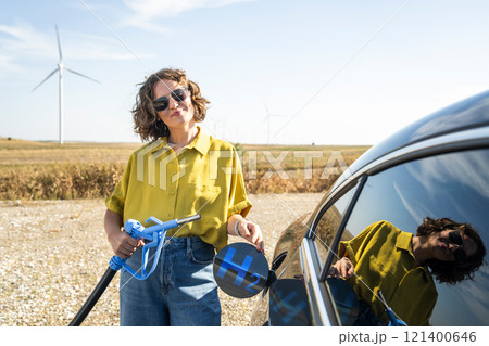 Woman holds a hydrogen fueling nozzle. Refueling car with hydrogen fuel. Wind turbines in the background. Woman holds a hydrogen fueling nozzle. Refueling car with hydrogen fuel. Wind turbines in the background. 121400646