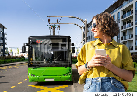 Woman with smartphone on the background of charging electric bus 121400652