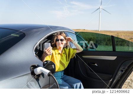 Woman with curve hairs wearing yellow shirt and sunglasses sits into charging electric car and holds smartphone. Wind turbines in the background Woman with curve hairs wearing yellow shirt and sunglasses sits into charging electric car and holds smartphone. Wind turbines in the background 121400662