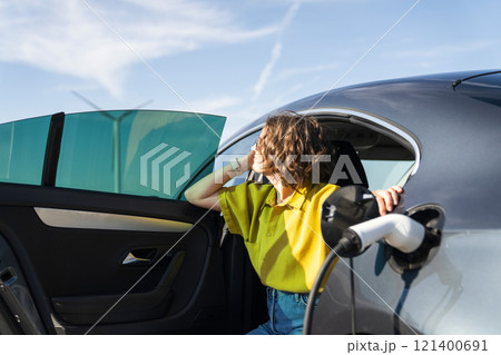 Woman with curve hairs wearing yellow shirt sits into charging electric car. Wind turbines in the background Woman with curve hairs wearing yellow shirt sits into charging electric car. Wind turbines in the background 121400691
