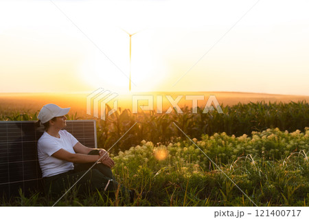 Woman wearing white cap sits next to solar panel at sunset. Wind turbines in the background Woman wearing white cap sits next to solar panel at sunset. Wind turbines in the background 121400717