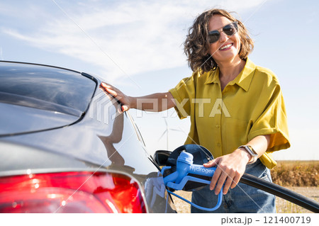 Woman with fueling hydrogen car. Refueling car with hydrogen fuel. Wind turbines in the background 121400719