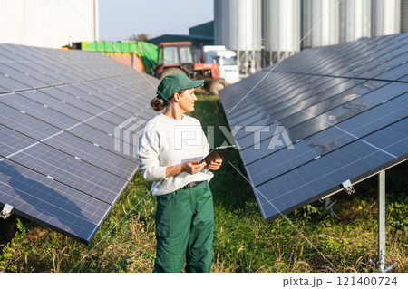 Female farmer with digital tablet on a modern farm using solar panels. Agricultural silos in the background. 121400724