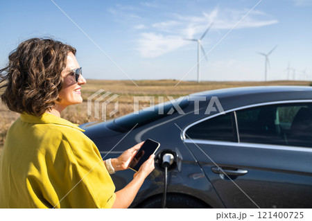 Woman with curve hairs wearing yellow shirt and sunglasses stands next to charging electric car and holds smartphone. Wind turbines in the background. Woman with curve hairs wearing yellow shirt and sunglasses stands next to charging electric car and holds smartphone. Wind turbines in the background. 121400725