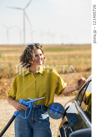 Woman holds a hydrogen fueling nozzle. Refueling car with hydrogen fuel. Wind turbines in the background Woman holds a hydrogen fueling nozzle. Refueling car with hydrogen fuel. Wind turbines in the background 121400726