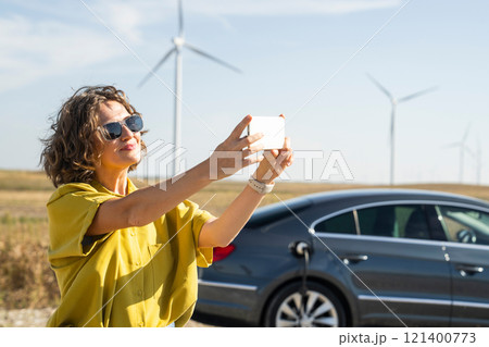 Woman with curve hairs wearing yellow shirt and sunglasses stands next to charging electric car and holds smartphone. Wind turbines in the background. Woman with curve hairs wearing yellow shirt and sunglasses stands next to charging electric car and holds smartphone. Wind turbines in the background. 121400773
