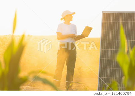 Woman farmer wearing white cap and t-shirt with laptop stands next to solar panel. Woman farmer wearing white cap and t-shirt with laptop stands next to solar panel. 121400794