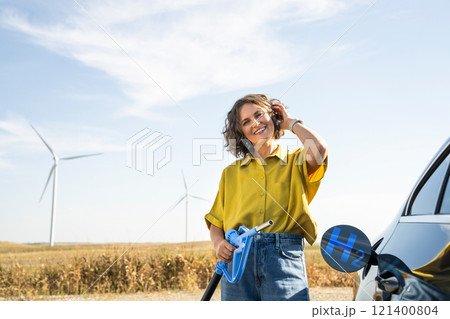Woman holds a hydrogen fueling nozzle. Refueling car with hydrogen fuel. Wind turbines in the background. Woman holds a hydrogen fueling nozzle. Refueling car with hydrogen fuel. Wind turbines in the background. 121400804