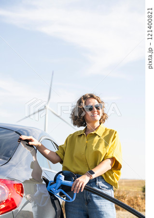 Woman with fueling hydrogen car. Refueling car with hydrogen fuel. Wind turbines in the background 121400821