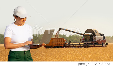 Female farmer with laptop on a background of harvesters. Smart farming concept 121400828
