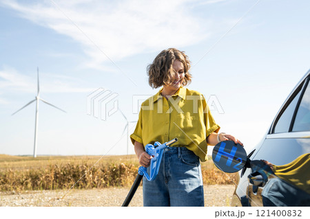 Woman holds a hydrogen fueling nozzle. Refueling car with hydrogen fuel. Wind turbines in the background. 121400832