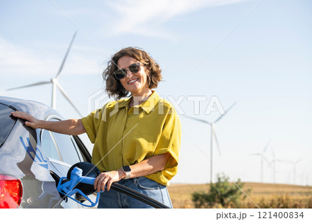 Woman holds a hydrogen fueling nozzle. Refueling car with hydrogen fuel. Wind turbines in the background. Woman holds a hydrogen fueling nozzle. Refueling car with hydrogen fuel. Wind turbines in the background. 121400834