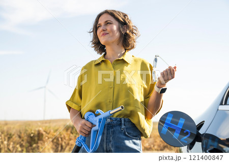 Woman holds a hydrogen fueling nozzle. Refueling car with hydrogen fuel. Wind turbines in the background. Woman holds a hydrogen fueling nozzle. Refueling car with hydrogen fuel. Wind turbines in the background. 121400847