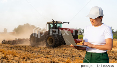 Female farmer with laptop controls an autonomous tractor on a smart farm Female farmer with laptop controls an autonomous tractor on a smart farm 121400930