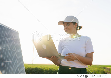 Woman farmer wearing white cap and t-shirt with laptop stands next to solar panel. Woman farmer wearing white cap and t-shirt with laptop stands next to solar panel. 121400936