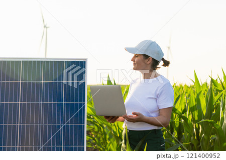 Woman farmer wearing white cap and t-shirt with laptop stands next to solar panel 121400952