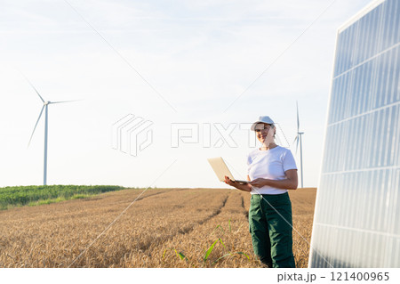 Woman farmer wearing white cap and t-shirt with laptop stands next to solar panel. Wind turbines in the background Woman farmer wearing white cap and t-shirt with laptop stands next to solar panel. Wind turbines in the background 121400965