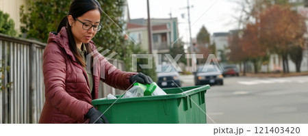 Woman putting recyclable items into green bin on residential street 121403420