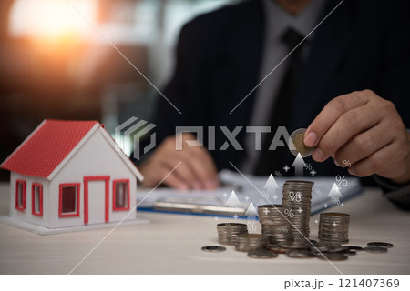 A person places a coin onto a stack near a model house, representing financial growth and real estate investment. 121407369