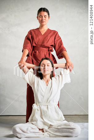 Masseuse making thai yoga massage. Woman therapist doing traditional massage treatment, stretching female patient's body. Client sitting on mat, enjoying hand massage in calm atmosphere. 121408944