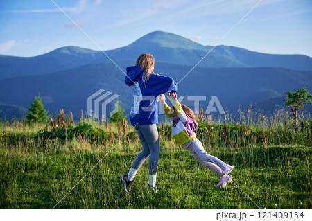 Young woman tourist swings, spending time together with little daughter on grassy hill, set against stunning mountain backdrop. Joyful moment captures essence of carefree fun and natural beauty. Young woman tourist swings, spending time together with little daughter on grassy hill, set against stunning mountain backdrop. Joyful moment captures essence of carefree fun and natural beauty. 121409134