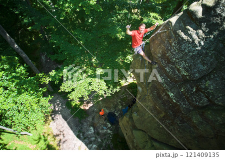 Aerial view of male rock climber ascending rugged limestone cliff with harness and rope for safety. Sportsman climbing on vertical large boulder at Dobvush Rocks in Carpathian mountains, Ukraine. Aerial view of male rock climber ascending rugged limestone cliff with harness and rope for safety. Sportsman climbing on vertical large boulder at Dobvush Rocks in Carpathian mountains, Ukraine. 121409135