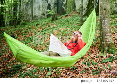 Man traveler in glasses relaxes in green hammock in lush forest. Male tourist examining map intently, surrounded by green foliage and tall rocks, planning outdoor adventure, hiking or climbing route. Man traveler in glasses relaxes in green hammock in lush forest. Male tourist examining map intently, surrounded by green foliage and tall rocks, planning outdoor adventure, hiking or climbing route. 121409137